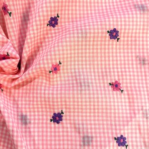 May include: Pink and white gingham fabric with embroidered flowers in shades of pink and purple. The flowers are small and scattered across the fabric. The fabric is slightly wrinkled, suggesting it is a piece of clothing or a textile.