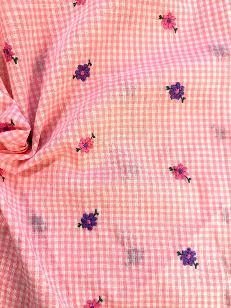 May include: Pink and white gingham fabric with embroidered flowers in shades of pink and purple. The flowers are small and scattered across the fabric. The fabric is slightly wrinkled, suggesting it is a piece of clothing or a textile.