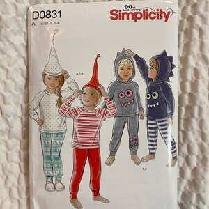 May include: Simplicity sewing pattern D0831 for children's pajamas. The pattern features various pajama designs, including a polka-dot top with deer-patterned pants, striped pajamas, and monster-themed hooded sets. The package is white with red and black text.