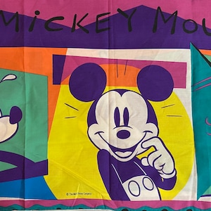 Könnte beinhalten: Ein farbenfrohes Mickey Mouse Bettlaken mit Mickey Mouse in drei verschiedenen Posen. Das Laken hat ein lebendiges Design mit den Worten "Mickey Mouse" oben. Der Hintergrund umfasst geometrische Formen in verschiedenen Farben, darunter Lila, Orange und Grün.