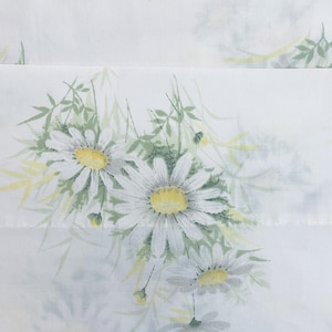 May include: White fabric featuring a daisy floral design. The daisies have white petals and yellow centres, with green leaves and stems. The fabric is folded, displaying the pattern in a repeating design.