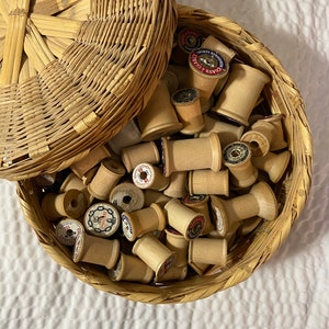 May include: A wicker basket filled with wooden spools of thread. The spools are wrapped in white thread and have various labels with red, blue, and gold lettering. The basket has a lid.