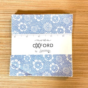 Peut inclure: Un ensemble de carrés de tissu avec un motif floral bleu et blanc. Le tissu est étiqueté "moda OXFORD by Sweetwater" et comprend une règle pour la mesure. Le tissu est sur une surface en bois.