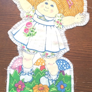 Puede incluir: Recorte de muñeca de tela que representa a una niña sonriente con coletas rubias atadas con lazos rosas. Lleva un vestido blanco con detalles florales y ribetes azules, y zapatos azules. La base muestra un campo verde con flores y setas.