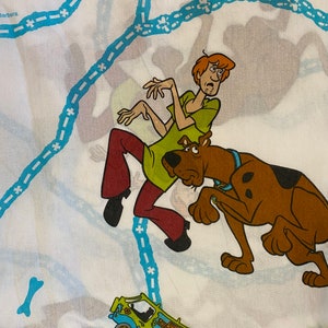 May include: White bed sheet featuring cartoon characters Shaggy and Scooby-Doo, along with the Mystery Machine van. Shaggy is wearing a green shirt and maroon pants. Scooby-Doo is brown with a blue collar. The van is green and blue.