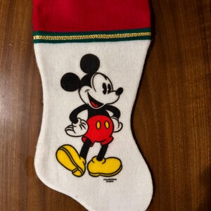 Puede incluir: Calcetín de Navidad blanco con un puño rojo y un ribete dorado. El calcetín presenta una imagen de dibujos animados de Mickey Mouse con sus característicos pantalones cortos rojos y zapatos amarillos.