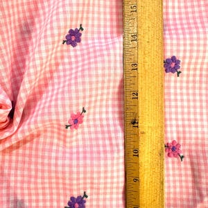May include: Pink and white gingham fabric with embroidered purple and pink flowers. A wooden ruler shows measurements in inches. The fabric is folded, showing the texture and pattern of the material.