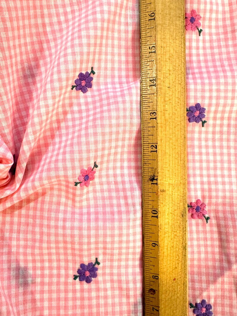 May include: Pink and white gingham fabric with embroidered purple and pink flowers. A wooden ruler shows measurements in inches. The fabric is folded, showing the texture and pattern of the material.