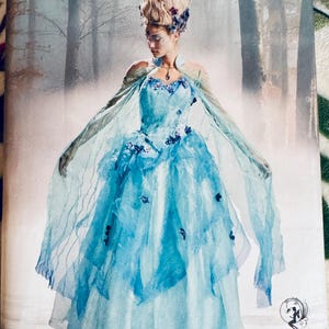 Puede incluir: El patrón Simplicity 8629 presenta un vestido de fantasía azul claro con mangas de capa transparentes y detalles florales. El patrón incluye tallas 6-14. La imagen también incluye el texto "90th anniversary Simplicity" y "Firefly Path."