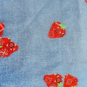 May include: A close-up of blue denim fabric featuring a repeating pattern of red strawberries. Each strawberry has a paisley design and green leaves. The fabric has a textured appearance.