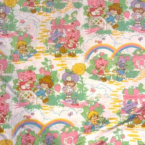 Op de afbeelding: Witte stof met een herhaald patroon van stripfiguren, aardbeien, regenbogen en bloemelementen. De personages zijn gekleed in verschillende outfits en zijn bezig met activiteiten zoals picknicken en spelen. Het kleurenpalet omvat roze, groen, geel en blauw.