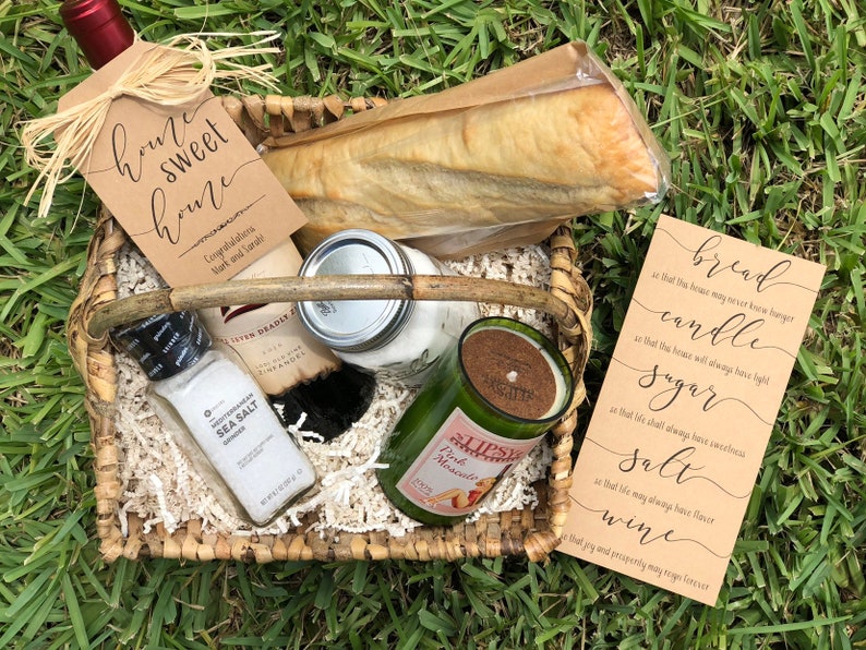 May include: A wicker basket filled with a housewarming gift set. The set includes a bottle of wine, a loaf of bread, a candle, sugar, salt, and a card with the text "bread, candle, sugar, salt, wine".
