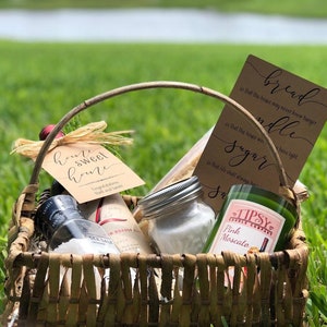 May include: A wicker basket filled with a housewarming gift set. The set includes a bottle of wine, a jar of sugar, a candle, and a card that reads "bread, candle, sugar, so that this house may never know hunger, so that this house will always have light, so that life shall always have sweetness".