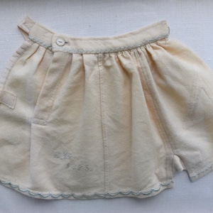 May include: Cream-colored vintage shorts with a button closure and scalloped trim. The shorts have pleats at the waist and a small pocket detail. Faint text is visible near the hem. A classic piece of antique clothing.