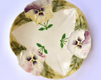 Pode incluir: Um prato de cerâmica na cor creme com formato triangular, apresentando três flores de amor-perfeito em relevo nos tons amarelo, roxo e branco. As bordas do prato são decoradas com folhas verdes e o centro é liso e simples.