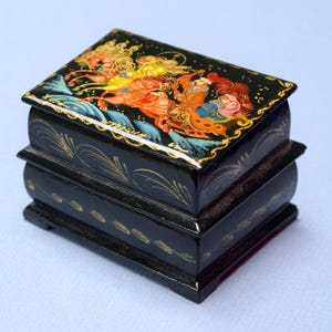 May include: Black lacquered box with a hinged lid, featuring a detailed, colorful painting of figures on horseback. The box has gold accents and a decorative design. The box is rectangular and has a layered design.
