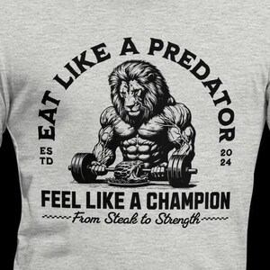 May include: A gray t-shirt with a black graphic of a lion lifting weights. The text "Eat Like A Predator" is in a circle around the lion. The text "Feel Like A Champion" is below the lion. The text "From Steak to Strength" is below the text "Feel Like A Champion".