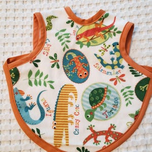 May include: White baby bib with an orange trim and backing. The bib features colorful illustrations of various reptiles, including a lizard, iguana, and tortoise, with the words "Silly", "Iguana", "Crazy Croc", and "Tortoise".