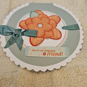 May include: A handmade greeting card with a scalloped edge. The card features a large orange flower with a white centre, surrounded by a light blue circle. A teal ribbon and bow accent the design, and the text "You're an amazing friend!" is printed on a small label.