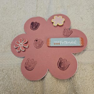 May include: A pink, flower-shaped craft with a white border. It features several small, stamped flower designs in a darker pink. The words "TRUE Friends!" are printed on a light blue rectangle, and there are two small, decorative flower accents.