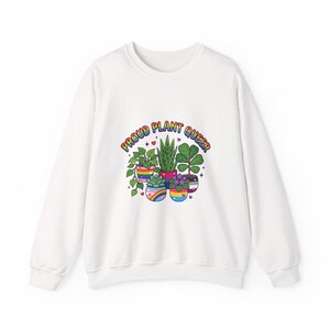 May include: A white long-sleeve sweatshirt featuring a graphic of five potted plants. The pots are decorated with various pride flag colors. Above the plants, the text reads PROUD PLANT QUEER in rainbow-colored letters, accented by small hearts.