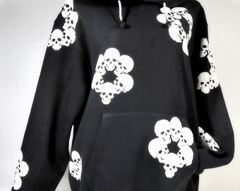 May include: A black hooded sweatshirt featuring a repeating white graphic pattern of circular skull clusters. The garment includes a front kangaroo pocket, long sleeves with ribbed cuffs, and a white striped hem. The hood has white drawstrings.