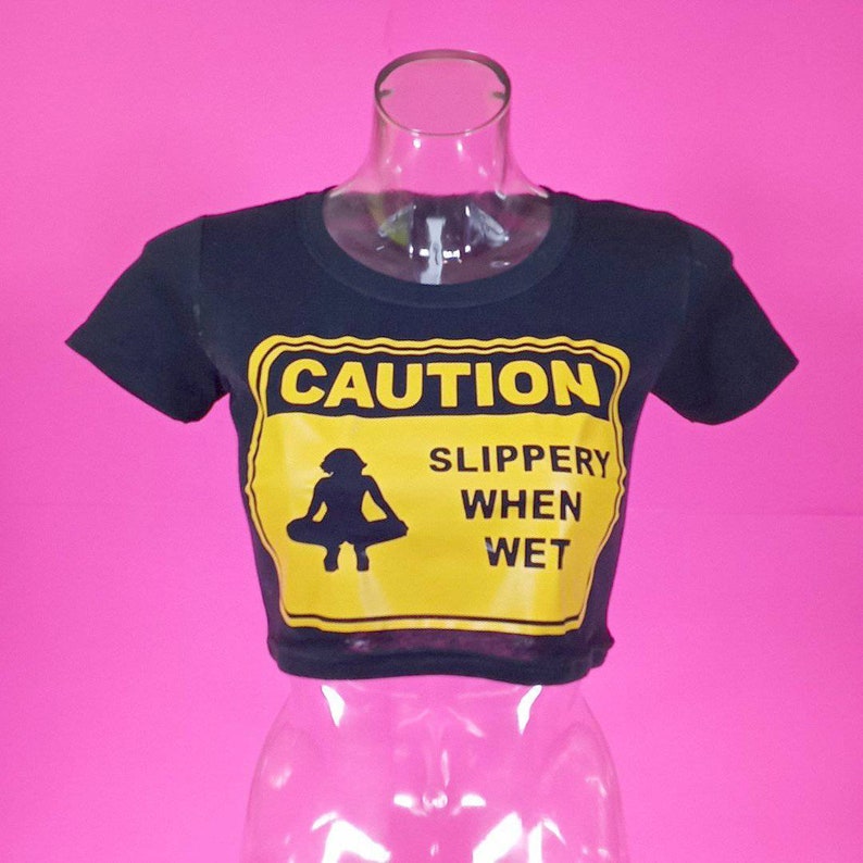 May include: Black cropped t-shirt with a yellow caution sign that says "CAUTION SLIPPERY WHEN WET" and a silhouette of a person in a yoga pose.