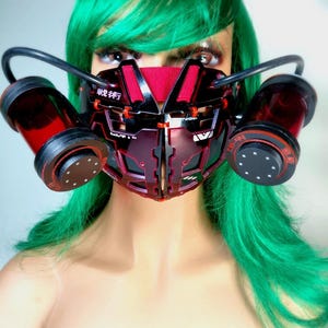 May include: A black and red futuristic mask with cylindrical attachments and the text "戦術". The mask has a geometric design with a red center panel. It is worn by a mannequin with green hair.