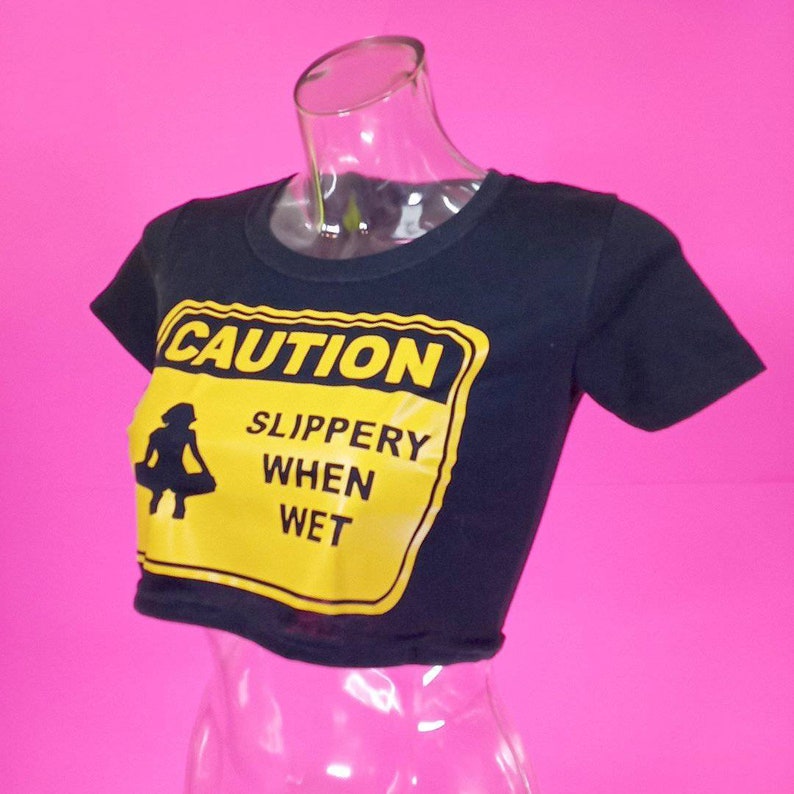 May include: Black cropped t-shirt with a yellow caution sign that says "CAUTION SLIPPERY WHEN WET". The sign has a silhouette of a person in a dress.