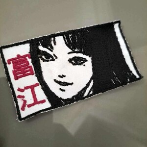 May include: White embroidered patch with a black and white image of a woman's face and the text "真紅" in red.