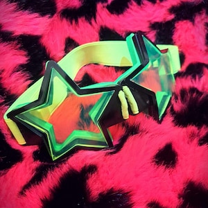 May include: Star-shaped neon green and black goggles with a yellow elastic strap. The goggles have a clear lens and are set against a vibrant pink and black patterned background.