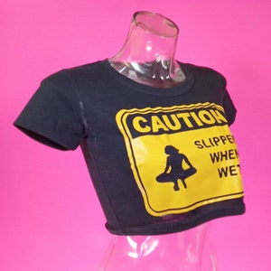 May include: Black cropped t-shirt with a yellow caution sign graphic that says "CAUTION SLIPPER WHEN WET" and a silhouette of a person sitting on a flying saucer.