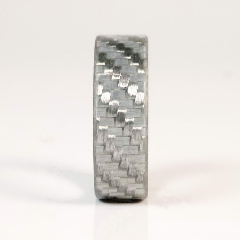 May include: A close-up of a grey ring with a woven carbon fibre pattern. The ring has a smooth, polished finish and a modern, minimalist design. The ring is approximately 0.8 cm wide.
