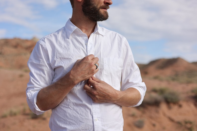 May include: A white long-sleeved button-down shirt. The shirt is being fastened. A black ring is visible on the person's finger. The background shows a desert landscape with red rock formations and a blue sky.