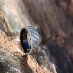 May include: A silver and black ring with a carbon fibre weave pattern is shown in close-up. The ring is set against a natural, textured rock background. The ring's interior is black.