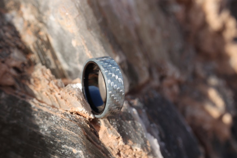 May include: A silver and black ring with a carbon fibre weave pattern is shown in close-up. The ring is set against a natural, textured rock background. The ring's interior is black.