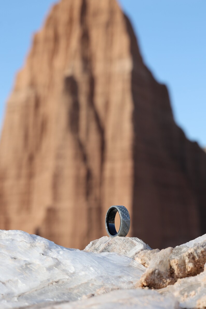 May include: A dark grey ring with a textured surface, set against a blurred backdrop of a rock formation and a clear blue sky. The ring is a band style, showcasing a modern design.