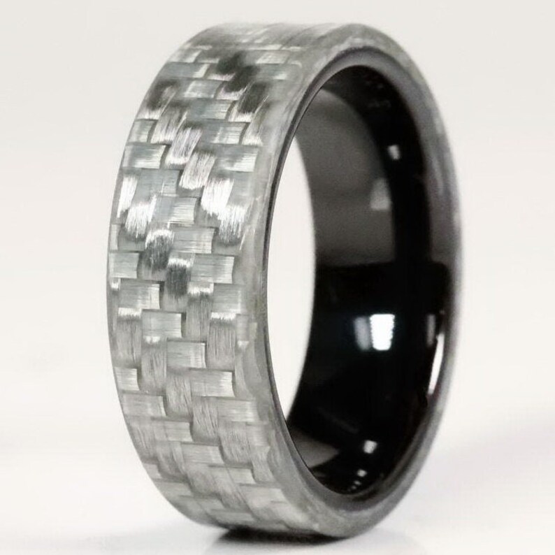 May include: A ring with a woven grey carbon fibre exterior and a glossy black interior. The ring has a smooth, rounded shape and a modern design. The carbon fibre pattern is clearly visible. The ring's width is approximately 0.8 cm.