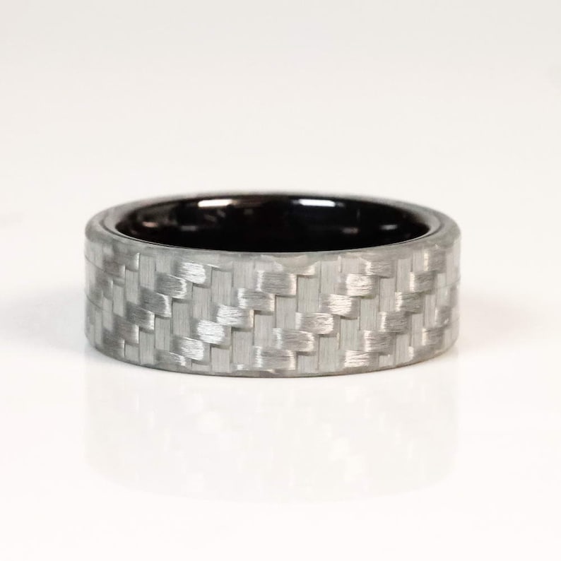 May include: A silver ring with a woven carbon fibre pattern on the outside and a black interior. The ring is a band style, with a smooth, polished finish. The ring is shown on a white background.