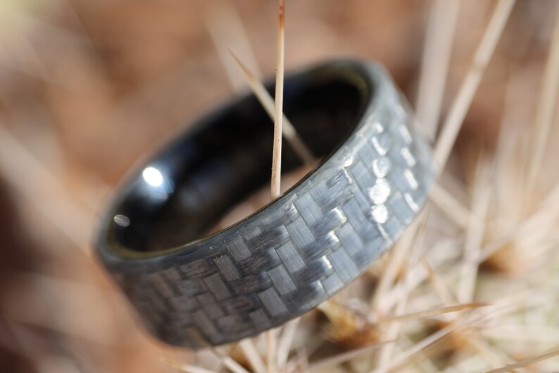 May include: A close-up shot of a dark grey ring with a woven carbon fibre pattern. The ring has a shiny, black interior. The ring's texture is visible against a blurred, natural background.