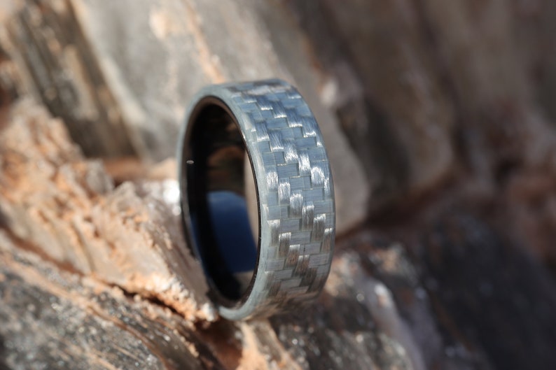May include: A close-up of a ring with a woven grey and black carbon fibre band. The ring has a black interior and is set against a blurred, natural background of light brown and grey rock.