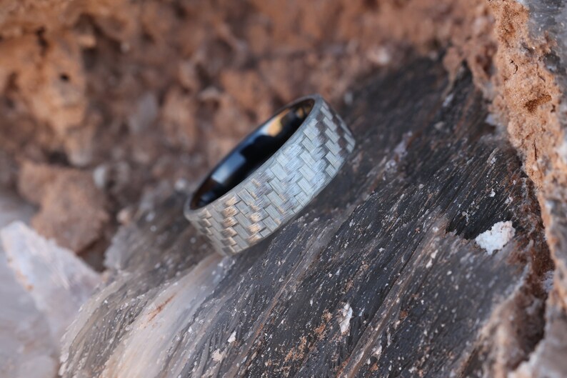 May include: A ring with a light grey woven pattern, set against a dark brown and grey stone surface. The ring has a dark interior. The design is modern and geometric. The ring's woven pattern is a key feature.
