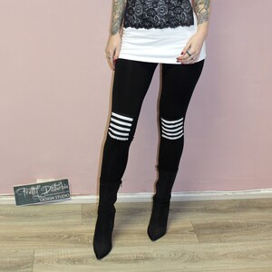 May include: A woman wearing a white tank top with a black lace overlay, black leggings with white stripes, and black boots. She is standing in front of a pink wall with a sign that says "Pretty Disturbia Design Studio".