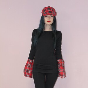 May include: A woman wearing a black long-sleeved shirt with red and black plaid cuffs and knee patches. She is also wearing a red and black plaid newsboy cap and black boots. The background is a light pink wall.