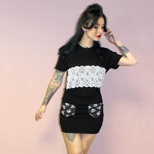 May include: A woman wearing a black t-shirt with a white lace overlay, a black mini skirt with white skull print, and black ankle boots. The skirt has a pocket on each side. The background is a light pink wall.