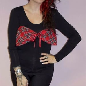 May include: A woman wearing a black long-sleeved shirt with a red and black plaid bow tied at the neckline. She has red and gray hair styled in a side part.
