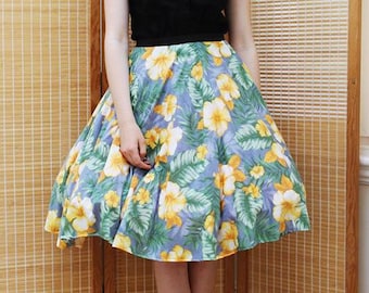May include: A woman wearing a black sleeveless shirt with a blue and white patterned collar, a floral print skirt, and black heeled sandals. She is standing in front of a wooden screen.