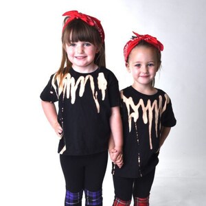 May include: Two young girls wearing black t-shirts with a bleach-splattered design and black leggings with plaid knee patches. Both girls are wearing red bandanas in their hair.