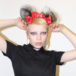 Puede incluir: Una persona con una camisa negra de botones y una falda de rayas blancas y negras. Lleva una diadema negra con lazos rojos y tul negro.