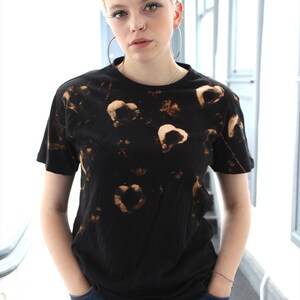 May include: A person wearing a black t-shirt with a bleach-dye design of brown hearts. The person is wearing blue jeans and has short blonde hair.